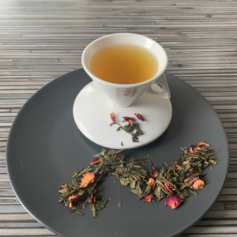 Grüner Tee Sencha Sakura Kirsche in weißer Tasse aufgegossen, lose grüne Teeblätter mit Blüten auf grauem Teller vor gestreiftem Holzhintergrund