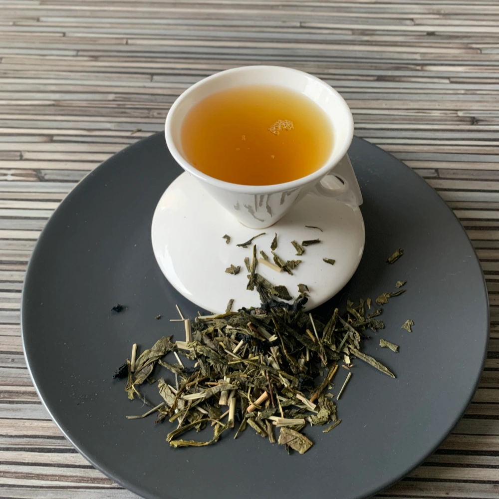 Grüner Tee Sencha Seealge Wakame Zitrus in weißer Tasse aufgegossen, lose grüne Teeblätter mit Algen und Kräutern auf grauem Teller vor gestreiftem Holzhintergrund