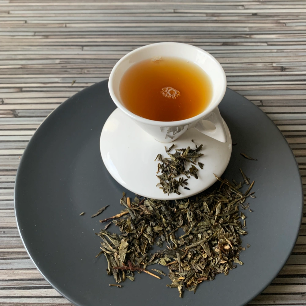 Grüner Tee Sencha Vanille in weißer Tasse aufgegossen, lose grüne Teeblätter auf grauem Teller vor gestreiftem Holzhintergrund