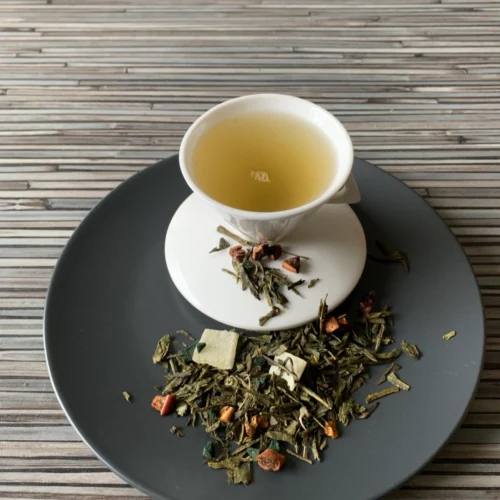 Grüner Tee Sencha Wunderbare Spirulina in weißer Tasse aufgegossen, lose grüne Teeblätter mit Blüten und Fruchtstücken auf grauem Teller vor gestreiftem Holzhintergrund