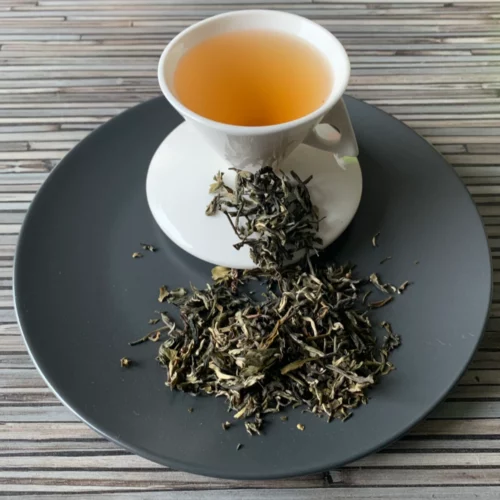 Grüner Tee China White Monkey in weißer Tasse aufgegossen, lose Teeblätter auf grauem Teller vor gestreiftem Holzhintergrund