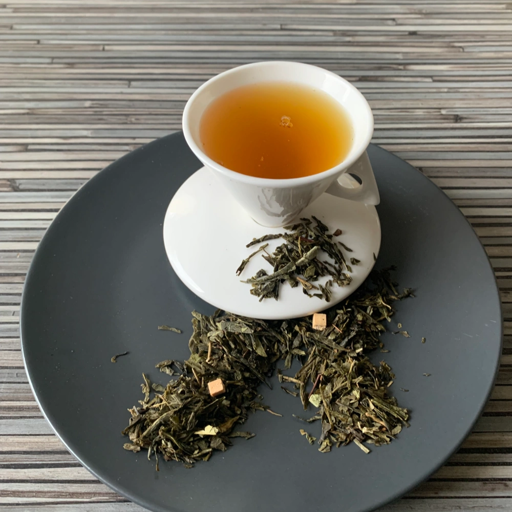 Grüner Tee Sencha Karamell Superior in weißer Tasse aufgegossen, lose grüne Teeblätter mit Karamellstücken auf grauem Teller vor gestreiftem Holzhintergrund
