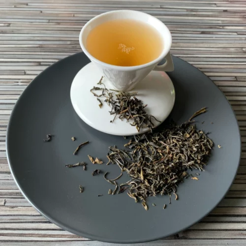 Grüner Tee Ceylon Idulgashinna in weißer Tasse aufgegossen, lose grüne Teeblätter auf hellem Teller vor gestreiftem Holzhintergrund