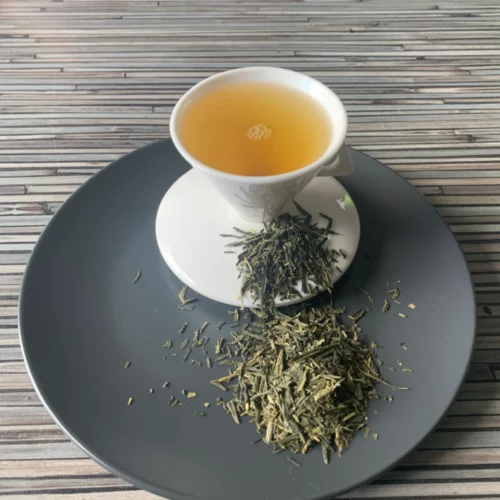 Grüner Tee Japan Bancha in weißer Tasse aufgegossen, lose Teeblätter auf grauem Teller vor gestreiftem Holzhintergrund