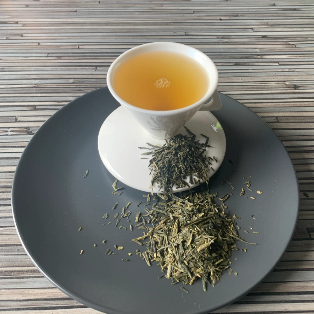 Grüner Tee Japan Bancha in weißer Tasse aufgegossen, lose Teeblätter auf grauem Teller vor gestreiftem Holzhintergrund