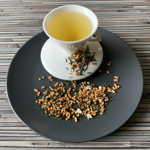 Grüner Tee Japan Genmaicha in weißer Tasse aufgegossen, lose Teeblätter mit Reis auf grauem Teller vor gestreiftem Holzhintergrund