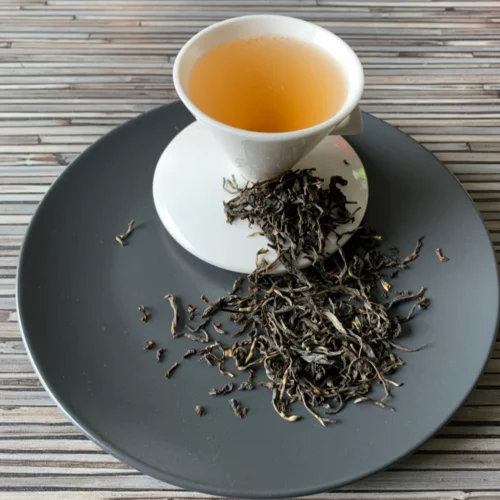 Grüner Tee Kolumbien Leafy Green in weißer Tasse aufgegossen, lose Teeblätter auf grauem Teller vor gestreiftem Holzhintergrund