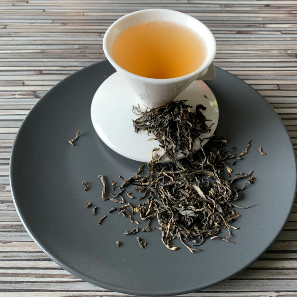 Grüner Tee Kolumbien Leafy Green in weißer Tasse aufgegossen, lose Teeblätter auf grauem Teller vor gestreiftem Holzhintergrund