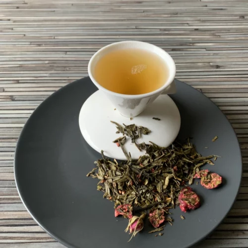 Grüner Tee Sencha Cranberry Rose in weißer Tasse aufgegossen, lose grüne Teeblätter mit Cranberries und Rosenknospen auf grauem Teller vor gestreiftem Holzhintergrund
