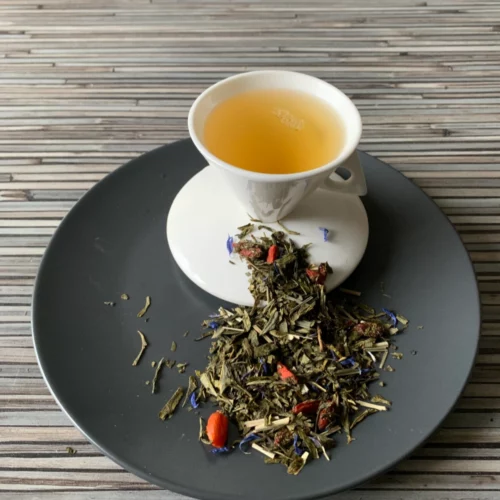 Grüner Tee Sencha Gojiberry Granatapfel in weißer Tasse aufgegossen, lose grüne Teeblätter mit Beeren und Blüten auf grauem Teller vor gestreiftem Holzhintergrund