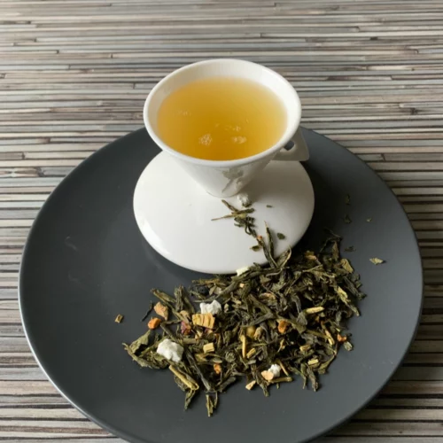 Grüner Tee Sencha Jet Set Tokio in weißer Tasse aufgegossen, lose grüne Teeblätter mit Fruchtstücken und Zitrusschalen auf grauem Teller vor gestreiftem Holzhintergrund