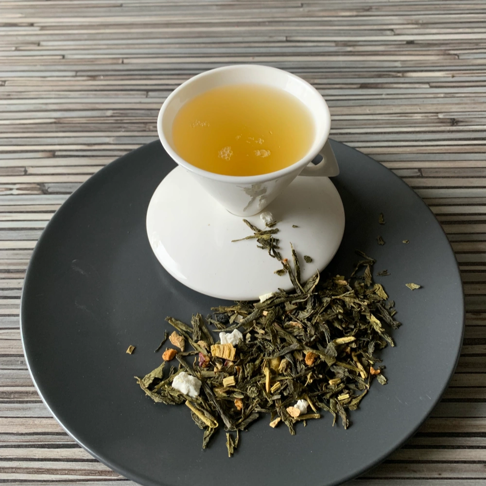 Grüner Tee Sencha Jet Set Tokio in weißer Tasse aufgegossen, lose grüne Teeblätter mit Fruchtstücken und Zitrusschalen auf grauem Teller vor gestreiftem Holzhintergrund
