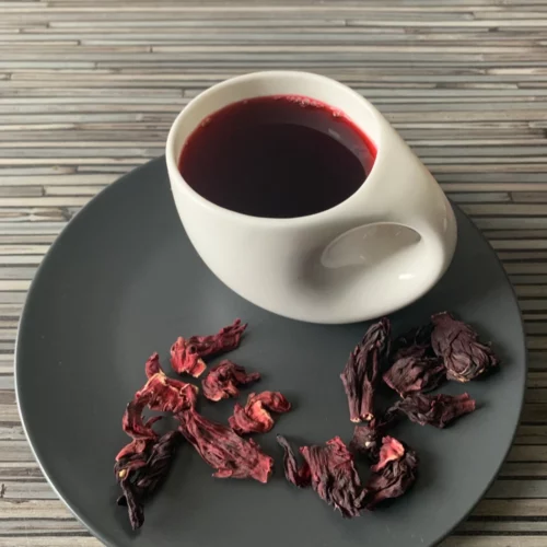 Hibiskusblüten Tee in weißer Tasse mit warmem Aufguss, auf grauem Teller mit losem Tee, arrangiert auf gestreiftem Holzhintergrund