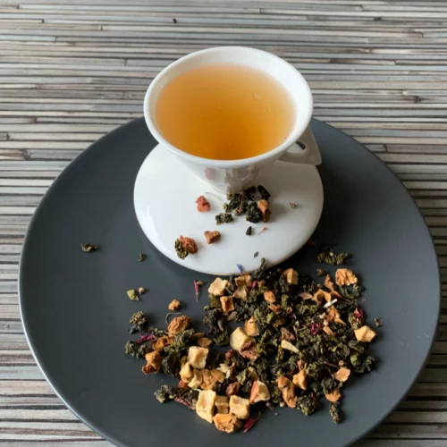 Holler die Holunderfee halbfermentiert mit Teeblättern, Apfelstücken, Heidekraut- und Kornblumenblüten und heller, klarer Tassenfarbe in weißer Teetasse auf gestreiftem Hintergrund