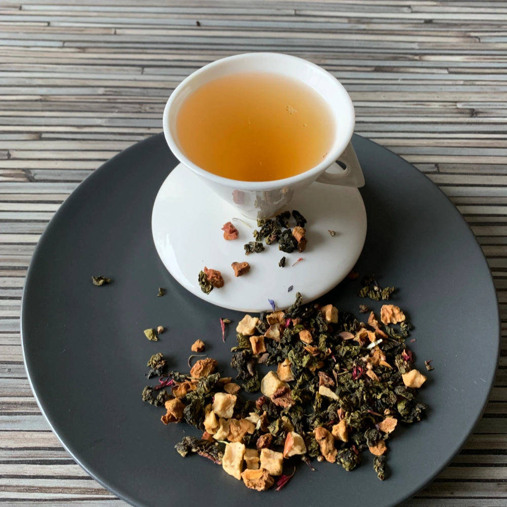 Holler die Holunderfee halbfermentiert mit Teeblättern, Apfelstücken, Heidekraut- und Kornblumenblüten und heller, klarer Tassenfarbe in weißer Teetasse auf gestreiftem Hintergrund