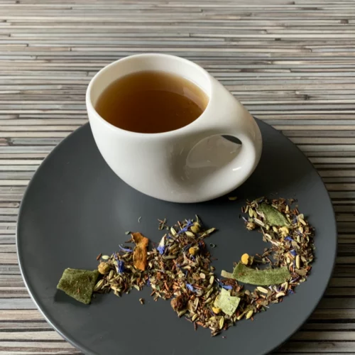 Grüner Tee Sencha Wunderbare Spirulina in weißer Tasse aufgegossen, lose grüne Teeblätter mit Blüten und Fruchtstücken auf grauem Teller vor gestreiftem Holzhintergrund