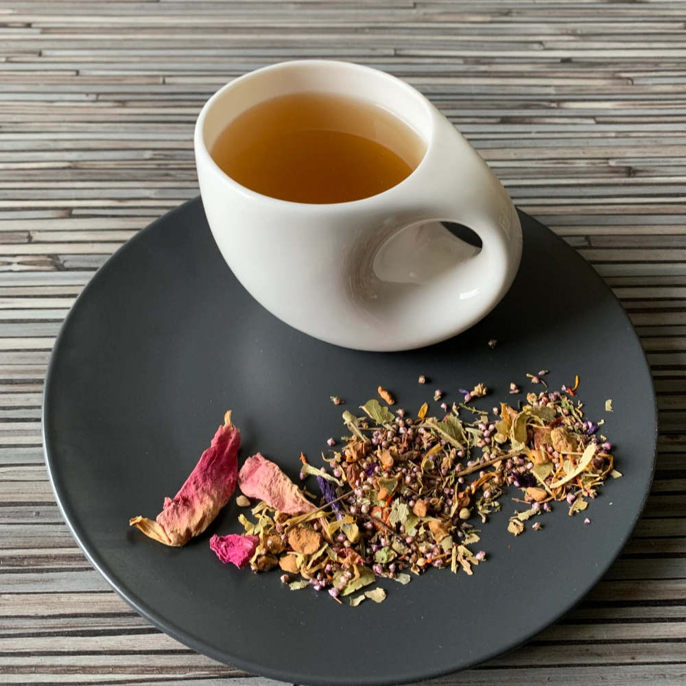 Kräutertee Blütenmeer Kirsch Himbeer in weißer Tasse aufgegossen, lose Blüten und Kräuter auf grauem Teller vor gestreiftem Holzhintergrund