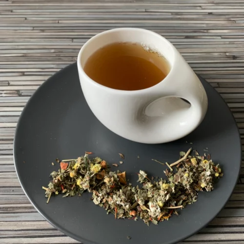 Kräutertee Familientee ohne Zusätze in weißer Tasse aufgegossen, lose Kräuter auf grauem Teller vor gestreiftem Holzhintergrund