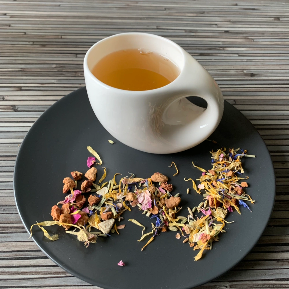 Kräutertee Farbenvielfalt ohne Aromazusätze in weißer Tasse aufgegossen, lose Teemischung auf grauem Teller vor gestreiftem Holzhintergrund