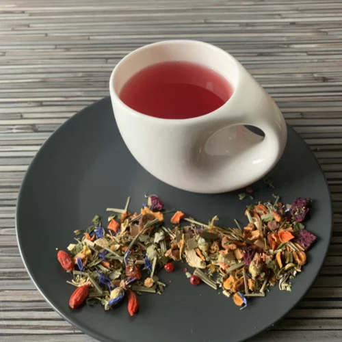 Kräutertee Feng Shui Tee Erdbeer Bergamotte in weißer Tasse aufgegossen, lose Kräuter und Blüten auf grauem Teller vor gestreiftem Holzhintergrund