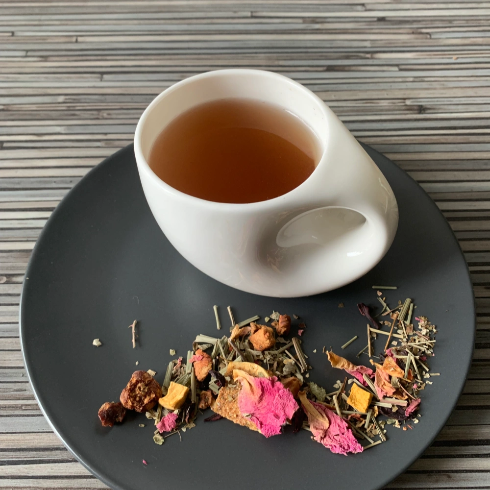 Kräutertee Goldener Herbst Kirsch Feigen Kürbis in weißer Tasse aufgegossen, lose Kräuter und Blüten auf grauem Teller vor gestreiftem Holzhintergrund