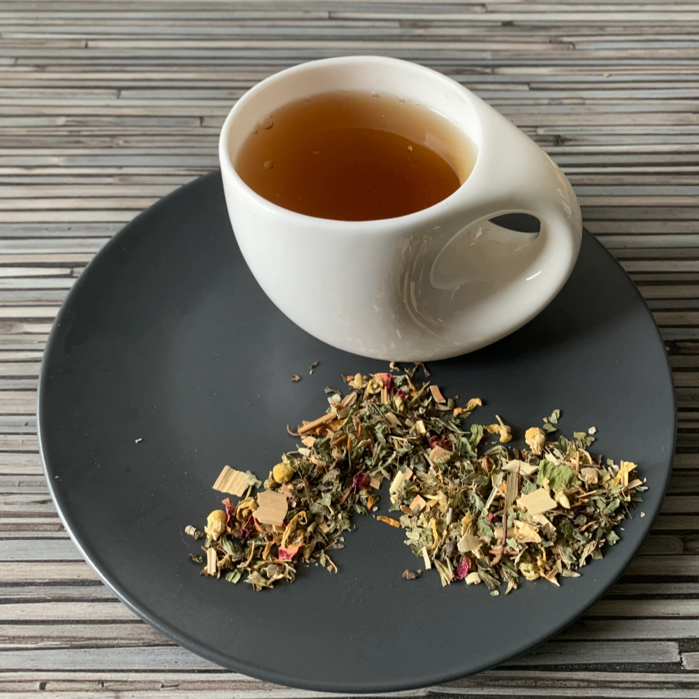 Kräutertee Heile Welt ohne Zusatz von Aromen in weißer Tasse aufgegossen, lose Kräuter und Blüten auf grauem Teller vor gestreiftem Holzhintergrund