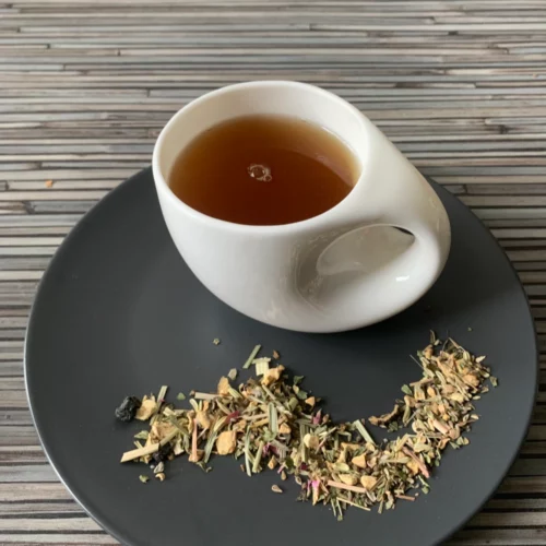 Kräutertee Ingwer Aronia ohne Aromazusatz in weißer Tasse aufgegossen, lose Kräuter und Blüten auf grauem Teller vor gestreiftem Holzhintergrund