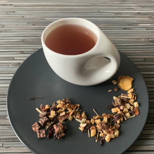 Kräutertee Ingwer Drachenfrucht Himbeere in weißer Tasse aufgegossen, lose Kräuter und Blüten auf grauem Teller vor gestreiftem Holzhintergrund