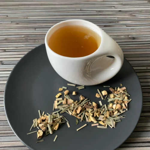 Kräutertee Ingwer Fresh ohne zusätzliche Aromen lose Kräuter und Blüten auf grauem Teller, aufgegossener Tee in weißer Tasse vor gestreiftem Holzhintergrund