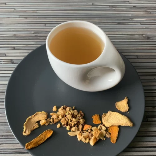 Kräutertee Ingwer Tonkabohne aufgegossen in heller Tasse, grobe Ingwer- und Gewürzstücke sichtbar, warmer Holzuntergrund