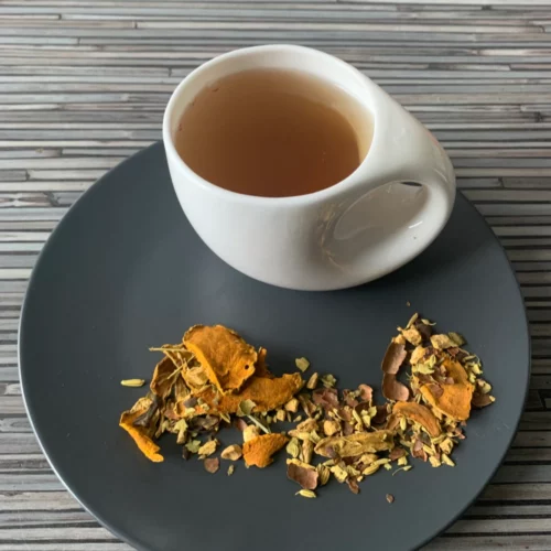 Kräutertee Kurkuma Gold mit Kakao aufgegossen in weißer Tasse, lose goldene Kurkuma- und Kakaomischung mit Ingwer, Fenchel, Anis und Vanille auf gestreiftem braun-dunkelbraunem Holzhintergrund