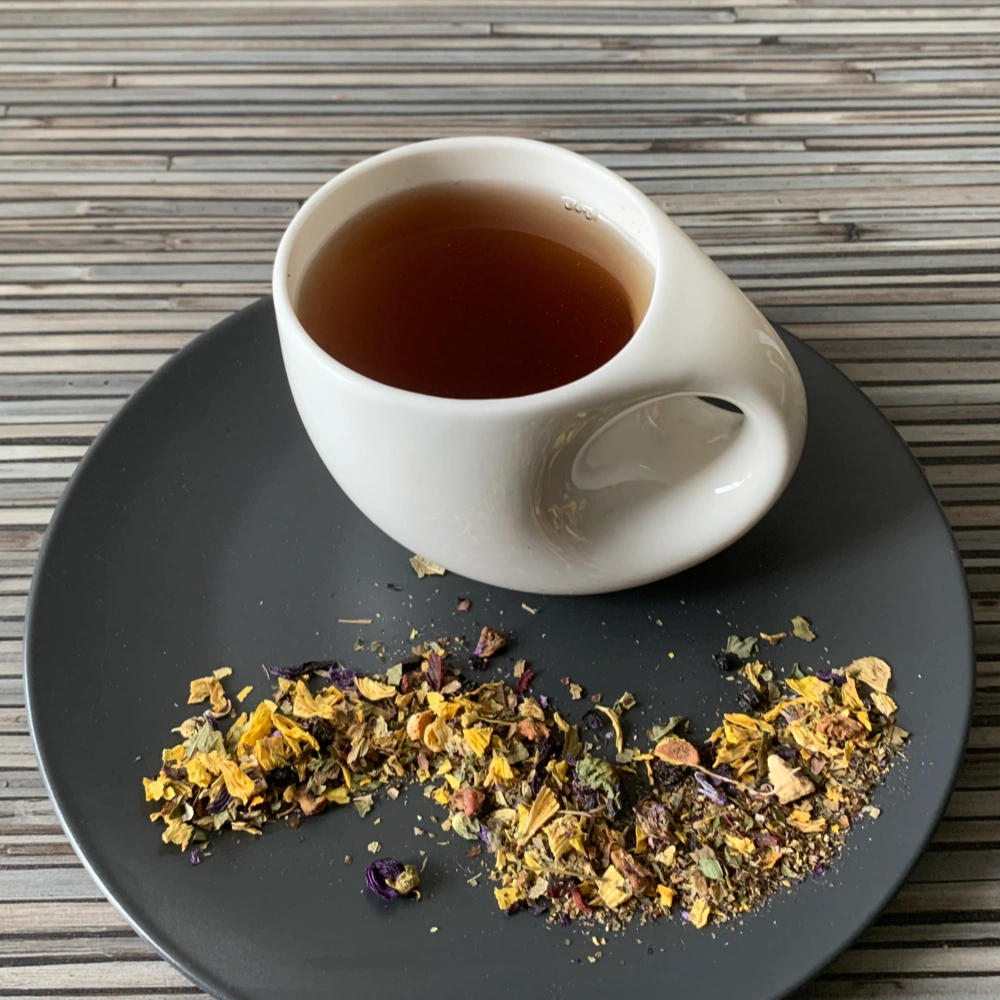 Kräutertee Ruhige Stunde in weißer Tasse aufgegossen, lose Kräuter und Blüten auf grauem Teller vor gestreiftem Holzhintergrund