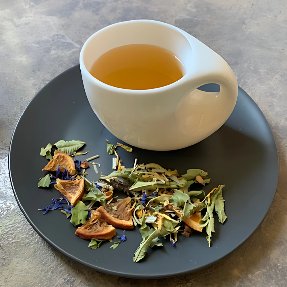 Kräutertee Sanfte Umarmung in weißer Tasse aufgegossen, lose Kräuter und Blüten auf grauem Teller vor gestreiftem Holzhintergrund