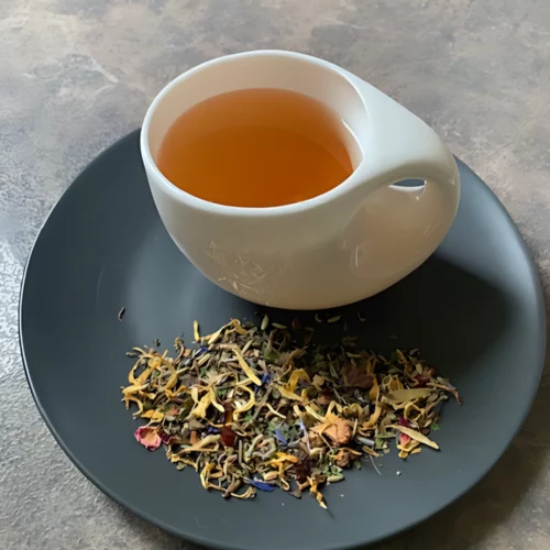 Kräutertee Schlummer Gut in weißer Tasse aufgegossen, lose Kräuter und Blüten auf grauem Teller vor gestreiftem Holzhintergrund