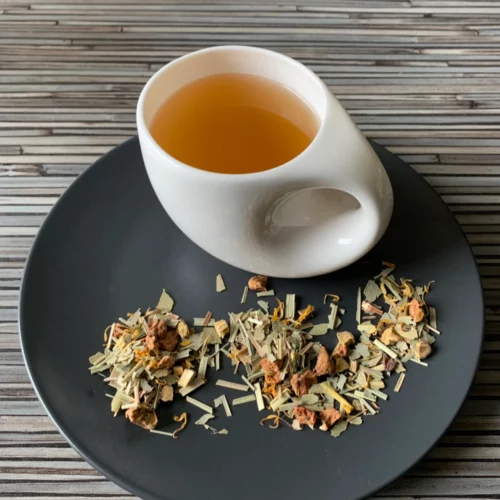 Kräutertee Seelenwohl ohne Zusatz von Aroma in weißer Tasse aufgegossen, lose Kräuter und Blüten auf grauem Teller vor gestreiftem Holzhintergrund