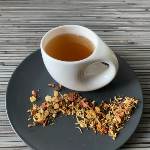 Kräutertee Sonnenkräuter in weißer Tasse aufgegossen, lose Kräuter und Früchte auf grauem Teller vor gestreiftem Holzhintergrund