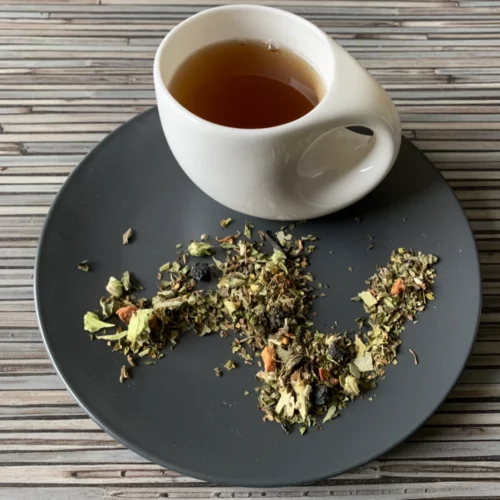 Kräutertee Super & Kühlend Minz Fichte Note in weißer Tasse aufgegossen, lose Kräuter, Aroniabeeren und Blätter auf grauem Teller vor gestreiftem Holzhintergrund