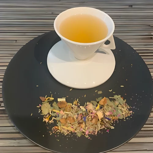 Kräutertee Urwaldfeeling Mangoblätter in weißer Tasse aufgegossen, lose Kräuter, Früchte und Mangoblätter auf grauem Teller vor gestreiftem Holzhintergrund