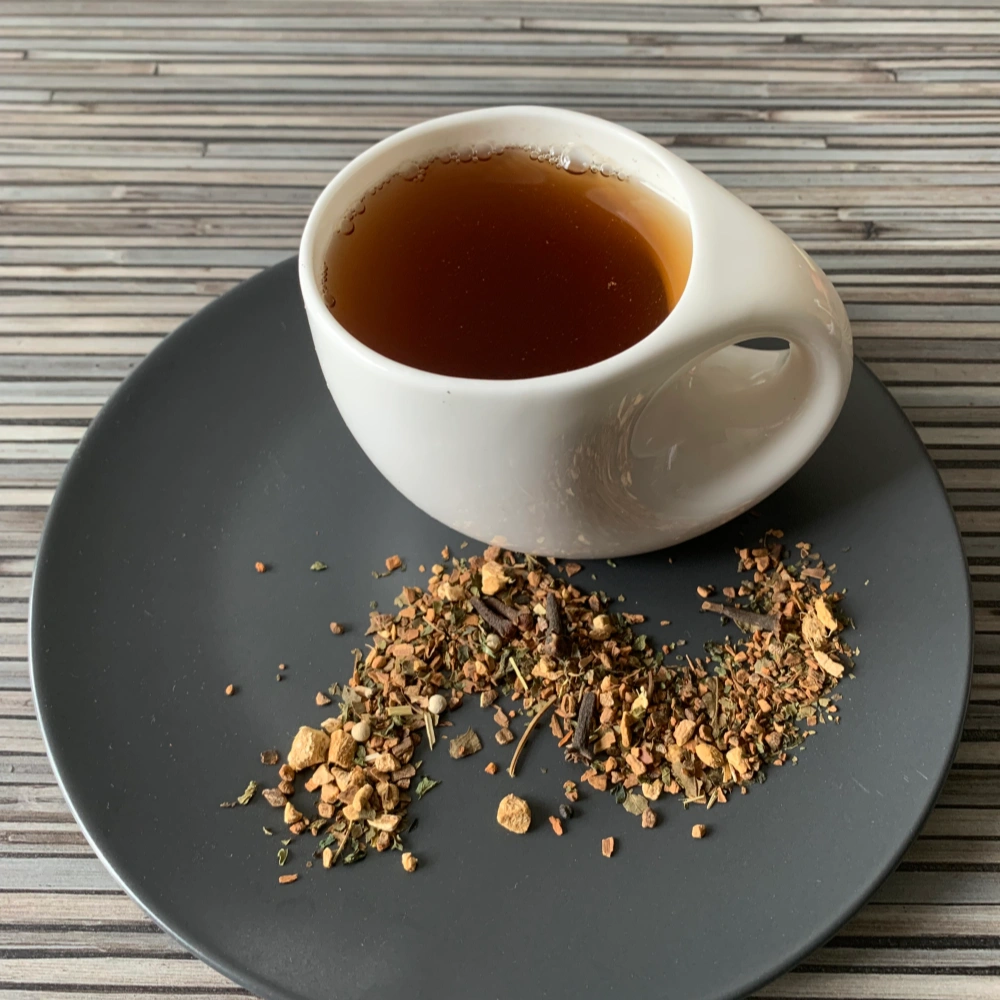Gewürztee Yoga Tee ohne Zusatz in weißer Tasse auf dunklem Teller auf gestreifter Holzplatte, loser Tee mit Gewürzen wie Zimt, Ingwer und Kardamom daneben