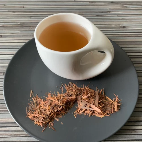 Lapacho Tajy Tee in weißer Tasse mit warmem Aufguss, auf grauem Teller mit losem Tee, arrangiert auf gestreiftem Holzhintergrund