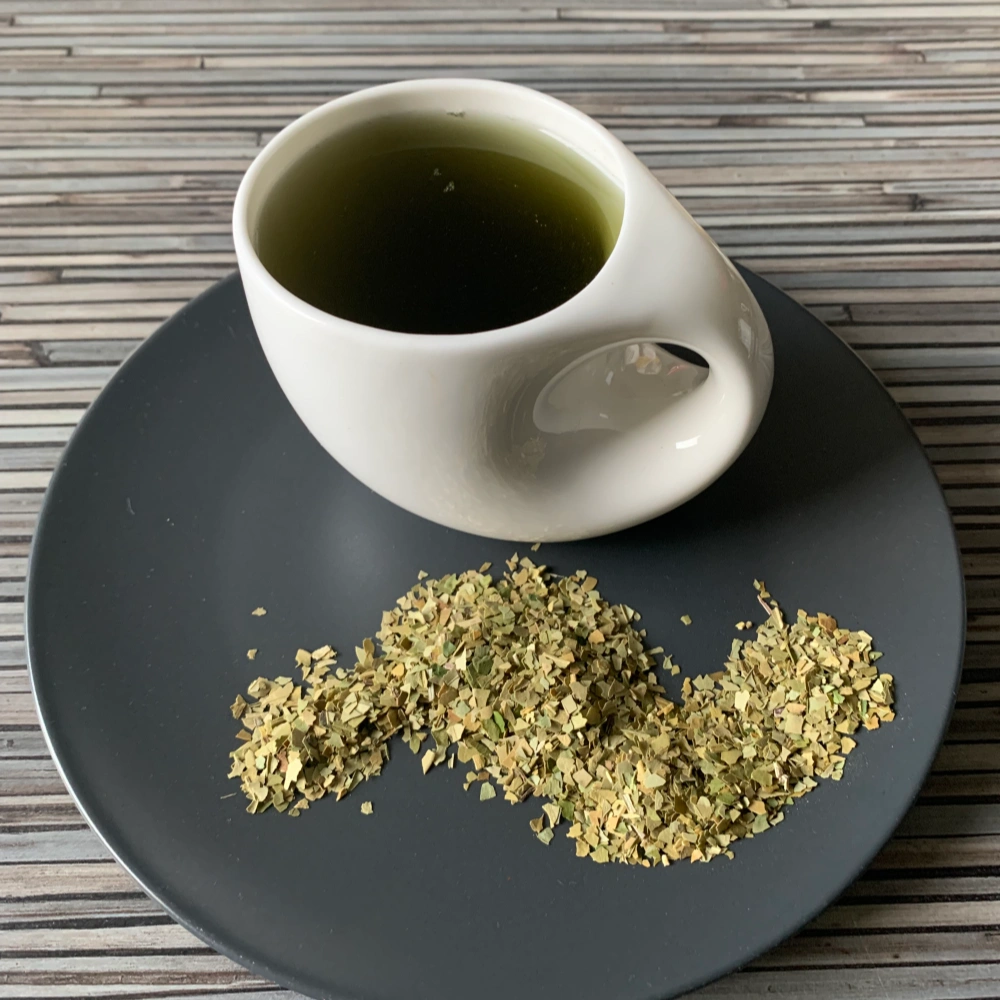 Mate Brasilien Yerba grün in weißer Tasse mit grünlichem Aufguss, lose Mateblätter auf grauem Teller, auf gestreiftem Holzhintergrund