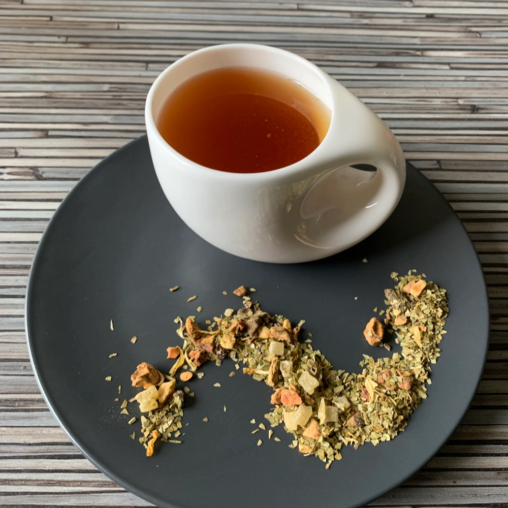 Kräutertee Mate Passionsfrucht aufgegossen in weißer Tasse, lose Mate-Mischung mit Passionsfrucht, Ananas und Ringelblumenblüten auf gestreiftem Holzhintergrund