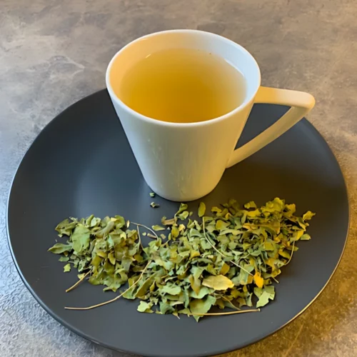 Moringablätter Tee in weißer Tasse mit hellgelben Aufguss, geschnittene Moringablätter auf grauem Teller, auf gestreiftem Holzhintergrund