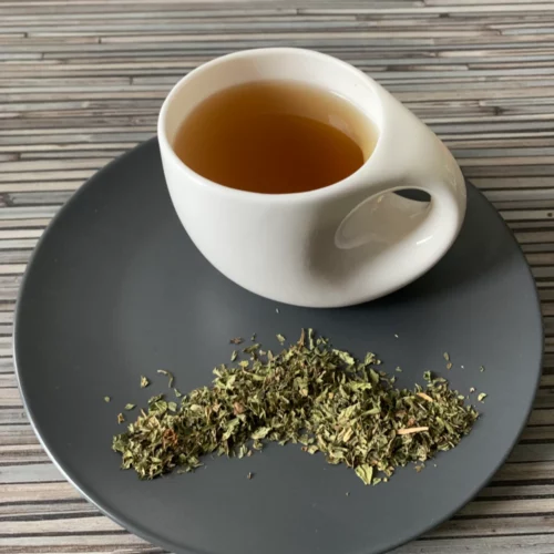 Nana Minze Spearmint geschnitten Tee in weißer Tasse mit warmem Aufguss, auf grauem Teller mit losem Tee, arrangiert auf gestreiftem Holzhintergrund