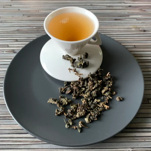 Oolong Auslese mit unterschiedlich gearbeiteten Oolong-Teeblättern und warmer bernsteinfarbener Tassenfarbe in weißer Teetasse auf gestreiftem Holzhintergrund
