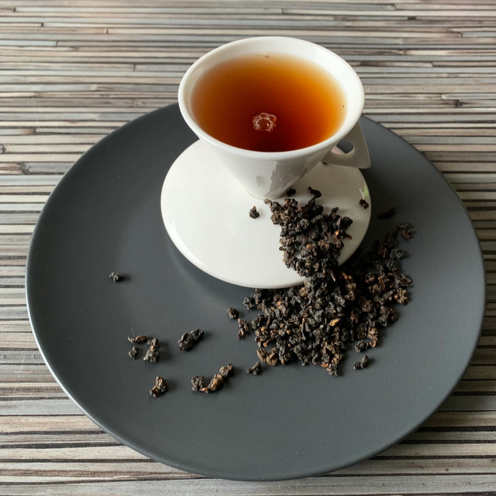 Formosa Dark Pearl Oolong mit dunkel gerollten Oolongperlen und bernsteinfarbener Tassenfarbe in weißer Teetasse auf gestreiftem Holzhintergrund