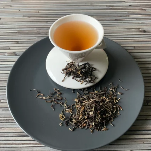 Oolong Gopaldhara Wonder Tea First Flush mit gedrehten Teeblättern, silbrig-weißen Blattspitzen und heller goldener Tassenfarbe in weißer Teetasse auf gestreiftem Holzhintergrund