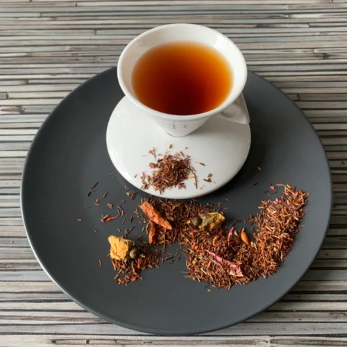 Rotbusch Feurige Schote Chili Kirsch mit Rooibosh und Chili-Schoten und kupferfarbener Tassenfarbe in weißer Teetasse auf gestreiftem Hintergrund