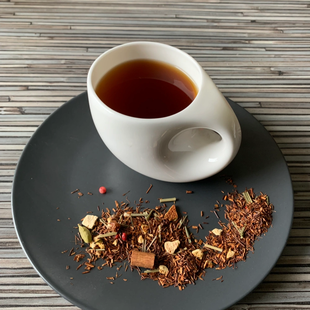 Rotbusch Gewürz Zitrone Pfeffer Note aromatisiert mit Rotbuschtee, Zitronengras und Gewürzen, kupferfarbene Tassenfarbe in weißer Teetasse auf gestreiftem Hintergrund