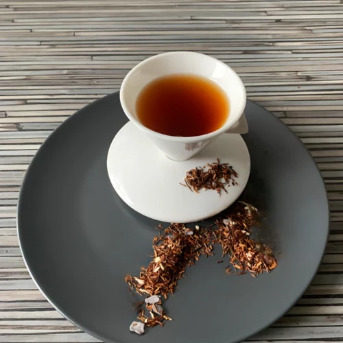 Rotbusch Hungerbändiger – kupferfarbener Rooibos mit Schokoladenplättchen in weißer Teetasse auf gestreiftem Hintergrund